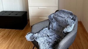 Um quarto com piso de madeira, uma poltrona cinza, uma cômoda branca com gavetas e um pequeno banquinho preto.