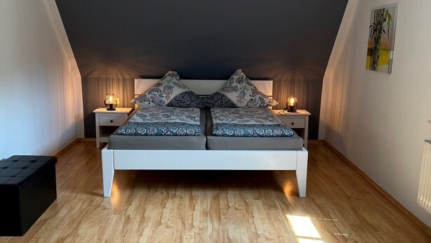 Quarto com teto inclinado, cama com roupa de cama estampada, duas luminárias de cabeceira e uma caixa de armazenamento preta.