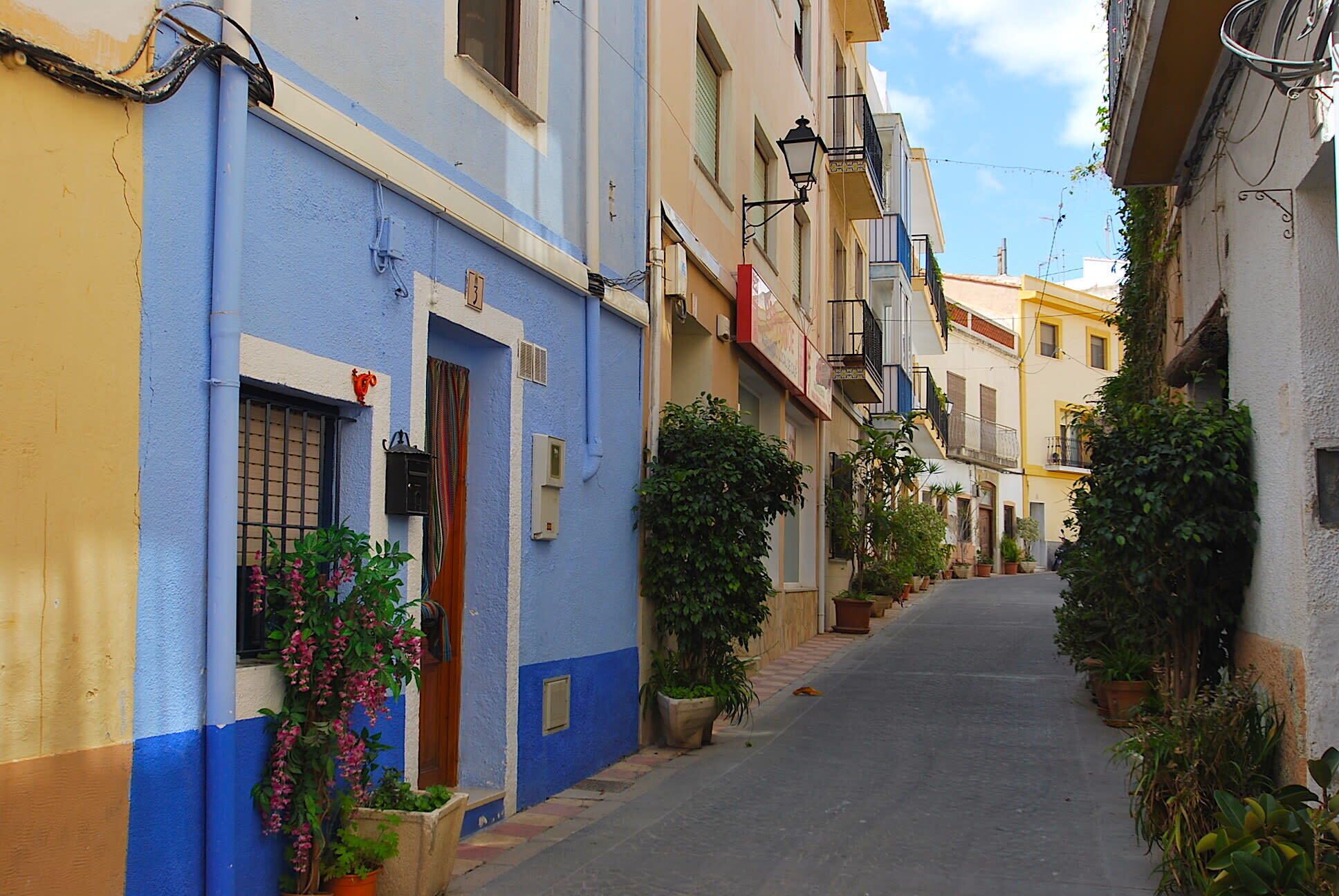 A narrow street lined with buildings, some with balconies and potted plants.