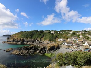 A coastal village with houses on a hillside, rocky shoreline, and clear blue water. - Louvain (Helston)