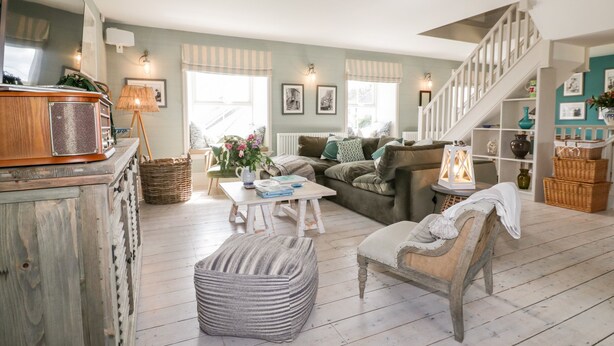 A cozy living room with a wooden coffee table, a striped ottoman, a green sofa, and a staircase leading to an upper level. - Louvain (Helston)