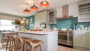 A modern kitchen with a central island, bar stools, stainless steel appliances, and teal backsplash. - Louvain (Helston)