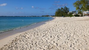 A wide sandy beach with clear blue water, a few people, and some trees.