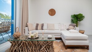 Een moderne woonkamer met een glazen salontafel, een witte bank met gestreepte kussens, een houten bijzettafeltje en een grote plant.