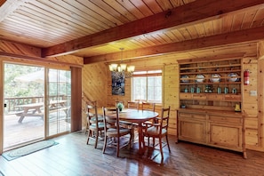 A wooden cabin interior with a dining table, chairs, a chandelier, and a kitchen with shelves.