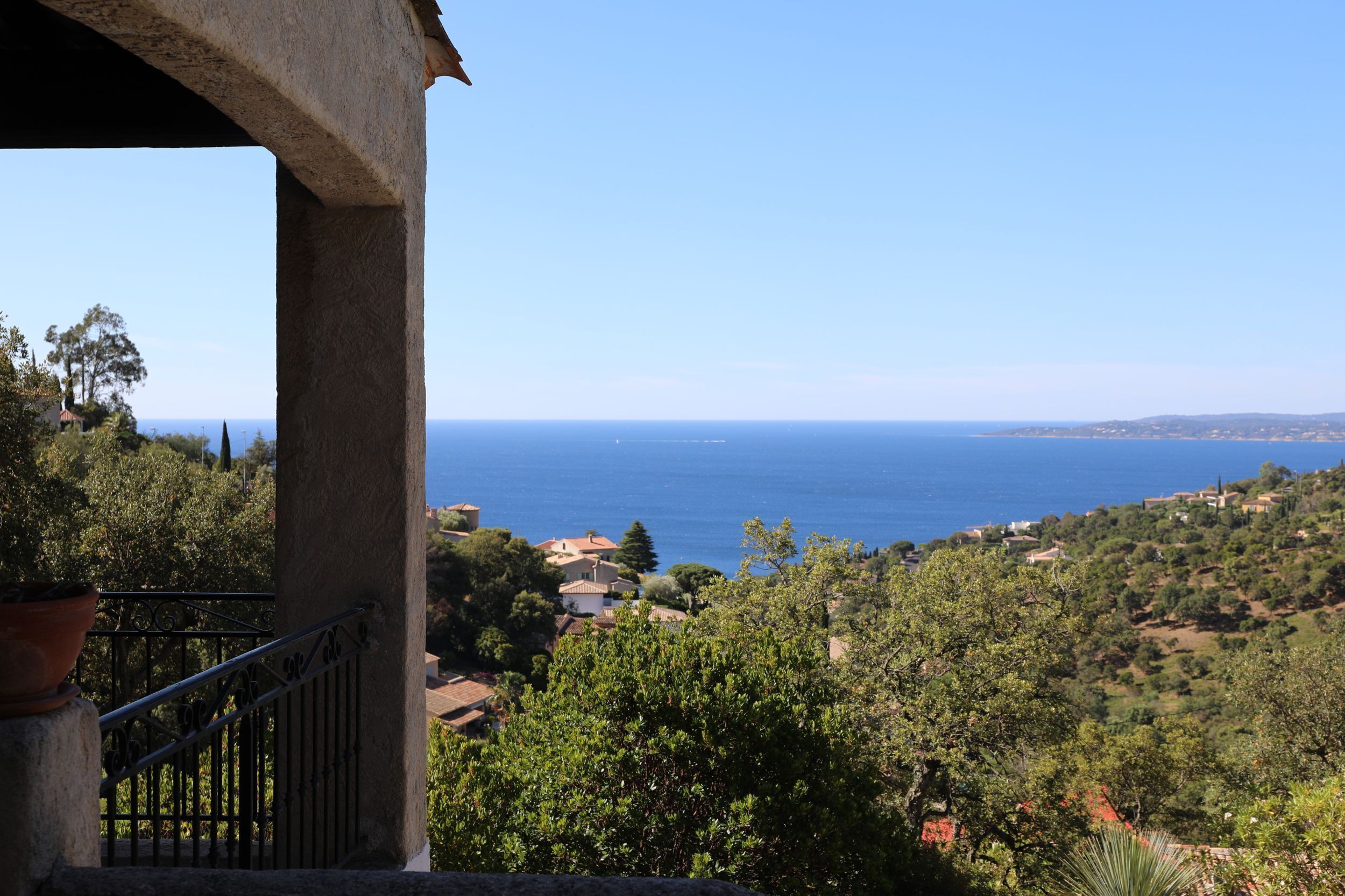 A coastal view from a balcony with a railing, overlooking a sea and a hilly landscape with buildings.