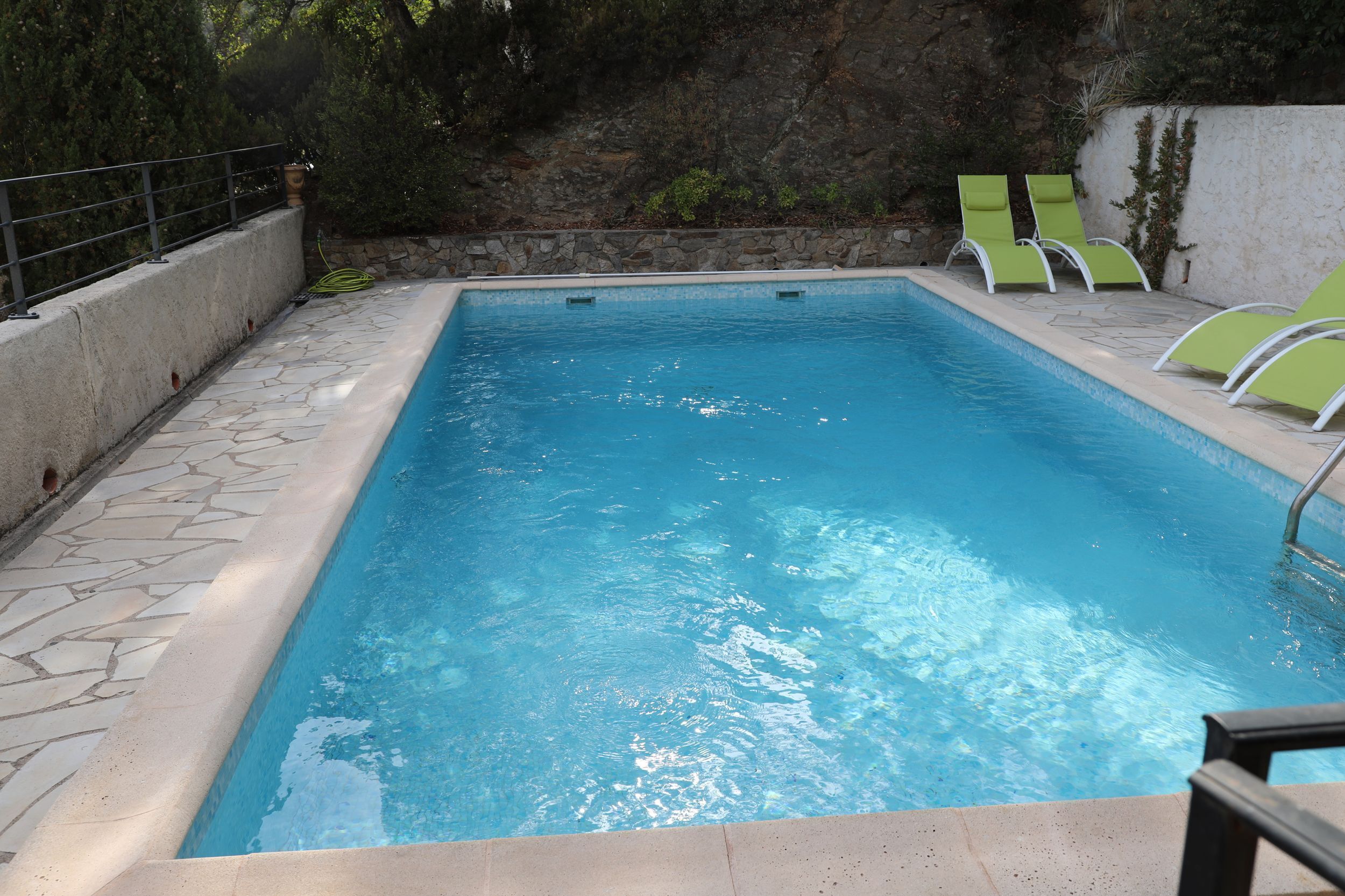 A rectangular swimming pool with clear blue water, surrounded by a stone-tiled deck and a low stone wall. There are two green lounge chairs on the right side of the pool.