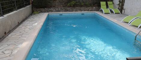 A rectangular swimming pool with clear blue water, surrounded by a stone-tiled deck and a low stone wall. There are two green lounge chairs on the right side of the pool.