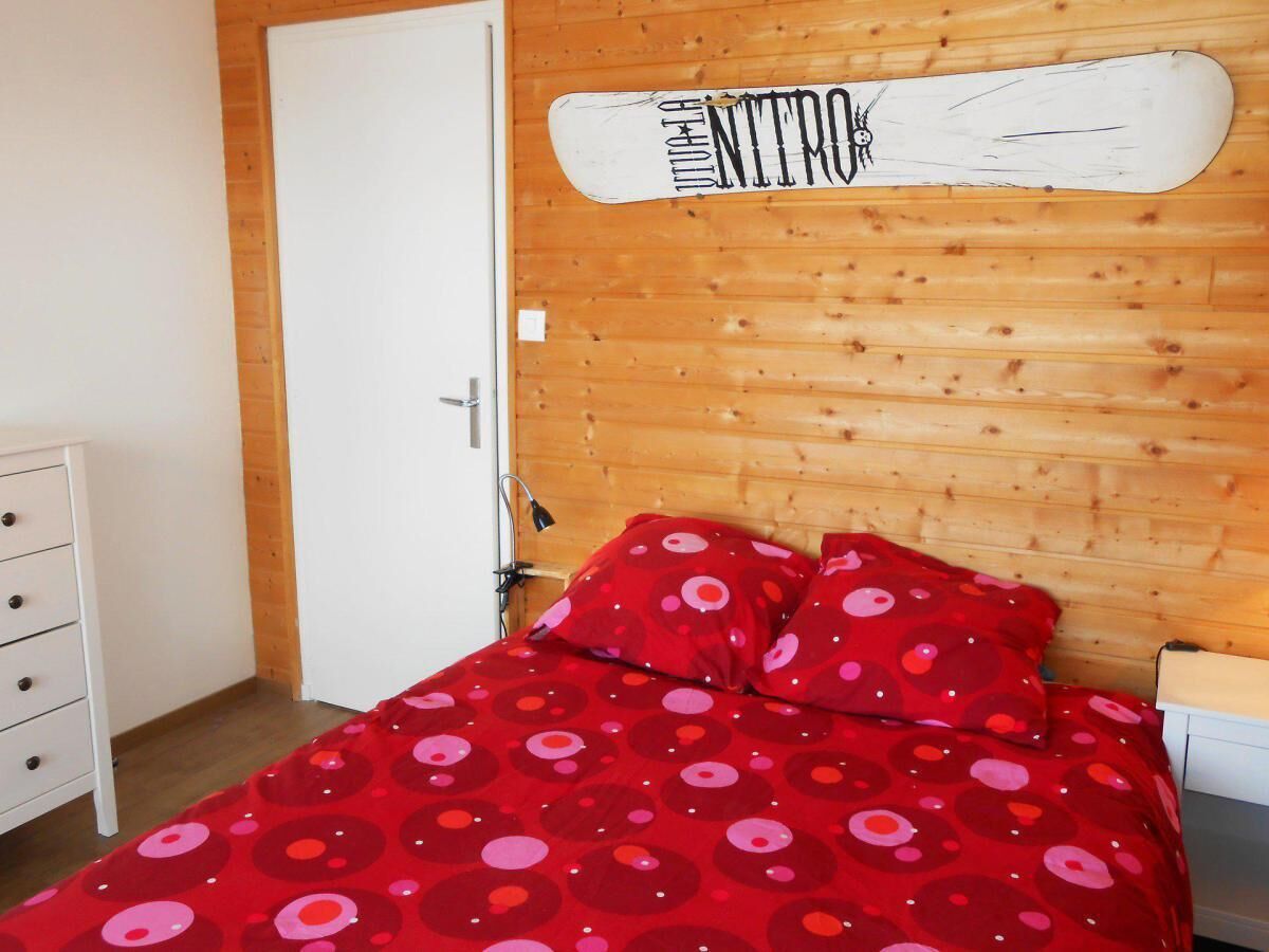 A bedroom with a wooden headboard, a bed with red bedding, a white dresser, and a white door.