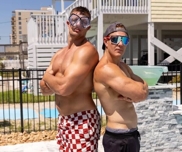 Two muscular men standing with their arms crossed in front of a pool area.