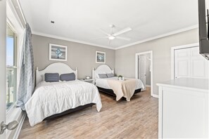 A bedroom with two beds, a ceiling fan, artwork on the walls, and a view of the outdoors. - 4202 Destination Sand Dollar - Oceanfront Pool (North Myrtle Beach)