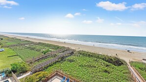 A beach view with a wooden boardwalk, grassy areas, and a clear sky.