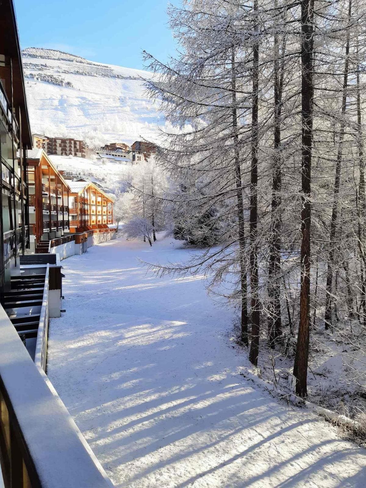 A snow-covered pathway flanked by buildings and trees, leading towards a mountainous backdrop.