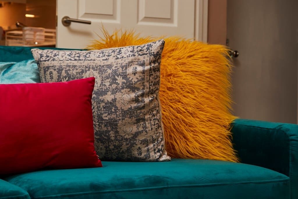 Un sofá color turquesa con tres cojines: uno rojo, uno estampado y uno amarillo mullido.