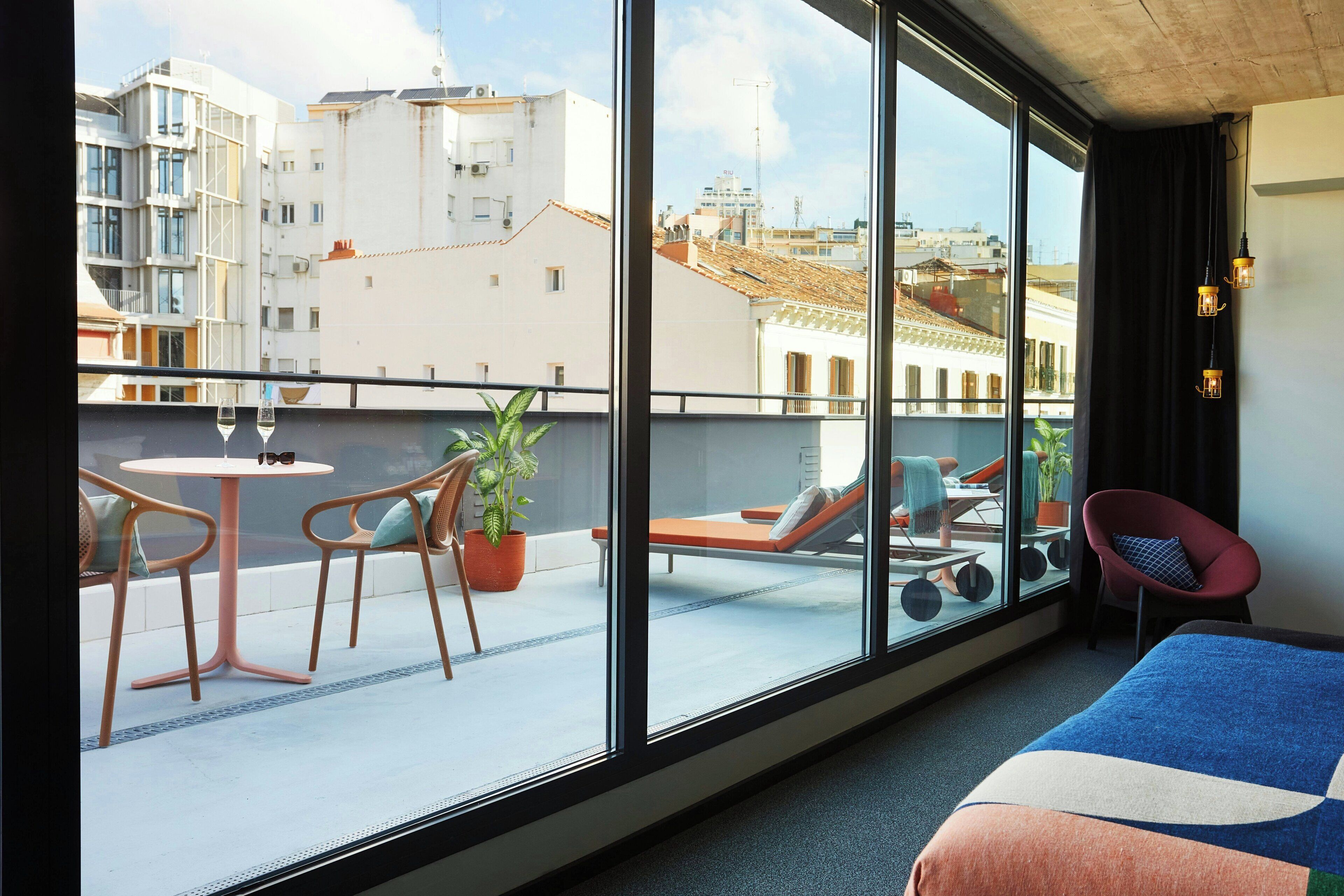 A hotel room with a large window, a bed, a small table, two chairs, a potted plant, and a balcony with a view of the city.