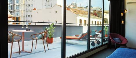 A hotel room with a large window, a bed, a small table, two chairs, a potted plant, and a balcony with a view of the city.