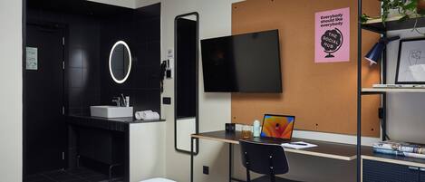 A modern hotel room with a bed, a desk with a laptop, a TV, a corkboard with a poster, and a bathroom with a mirror and sink.