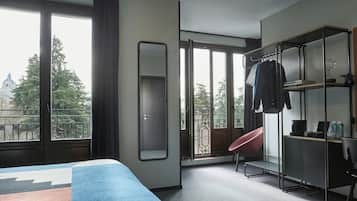 Hotelkamer met een bed, bureau, stoel, kledingkast en groot raam met uitzicht op bomen en een gebouw.