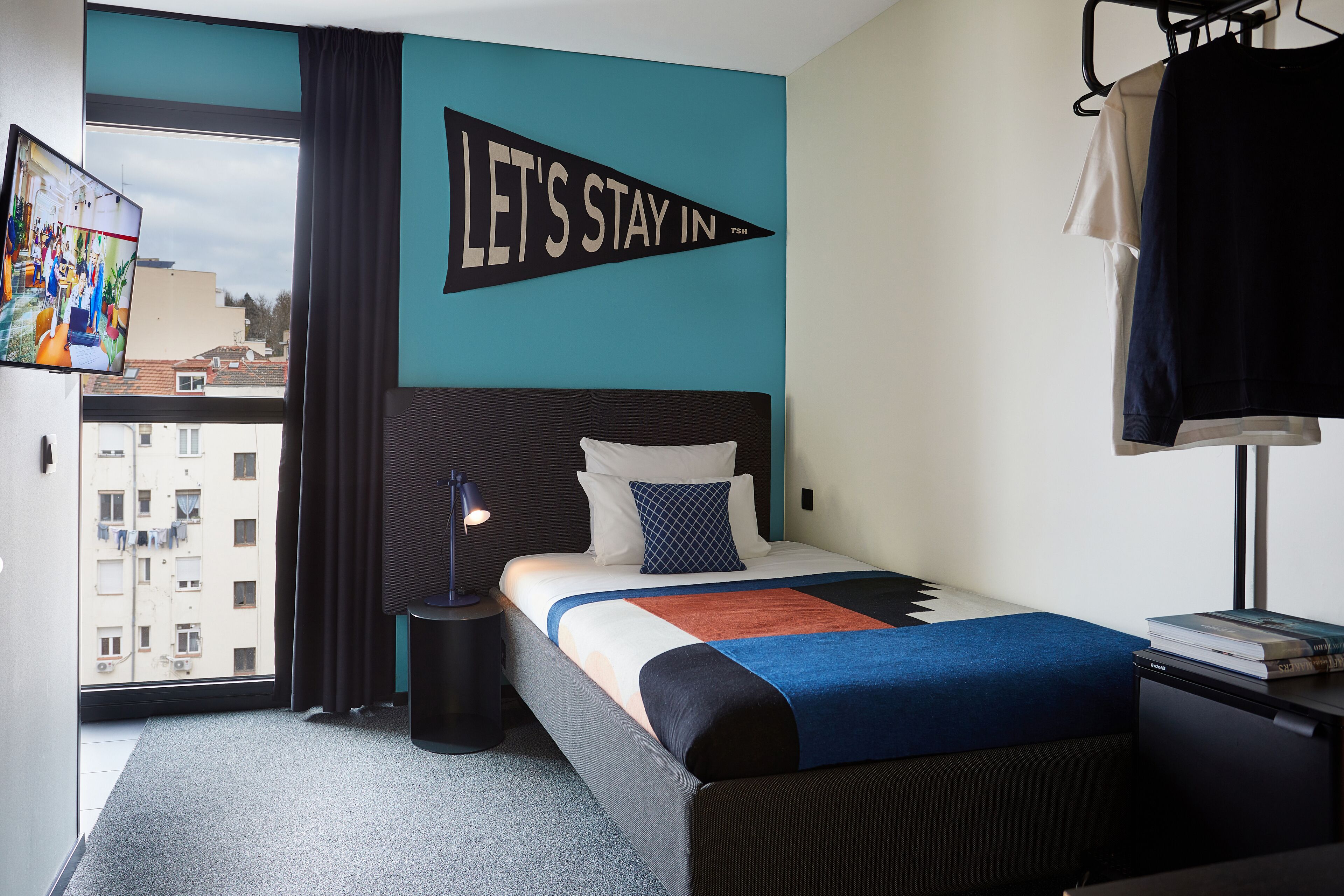 Una habitación de hotel con cama, televisor, ventana con cortinas y un cartel que dice 'LET'S STAY IN'.