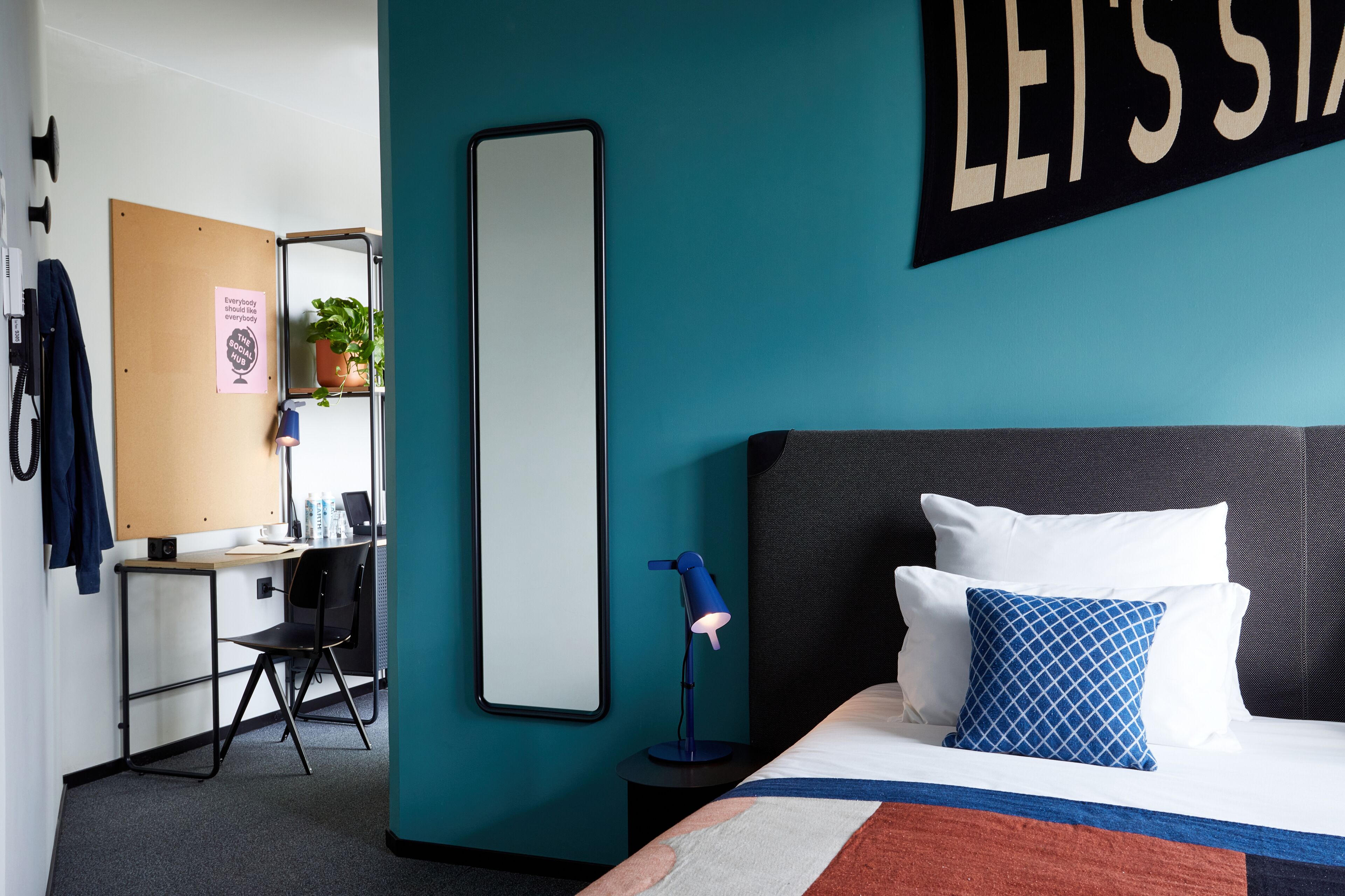 A modern hotel room with a teal accent wall, a large bed with white and blue patterned pillows, a desk with a chair, a mirror, and a hanging sign that reads 'LEISSIA'.