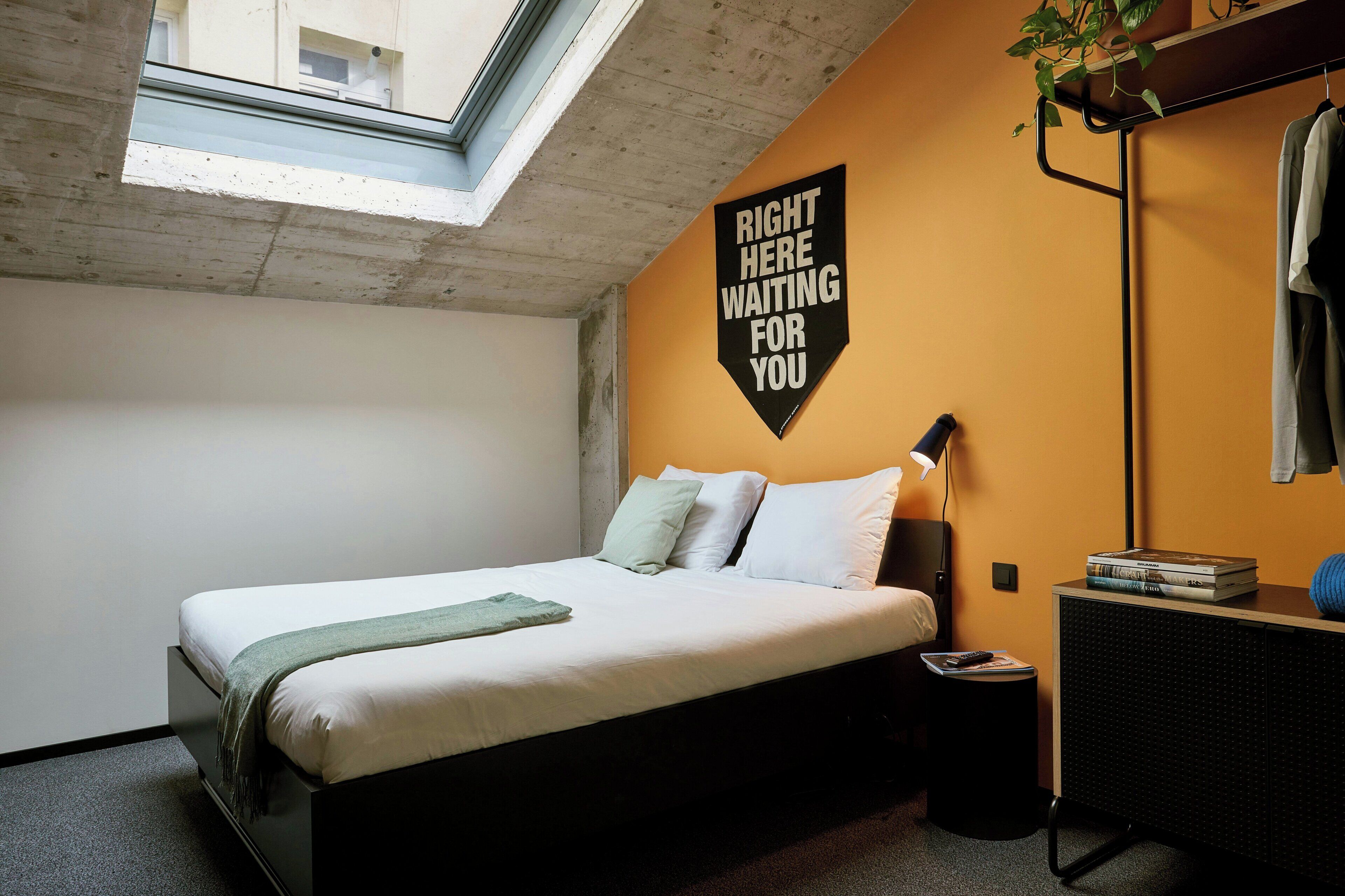 A bedroom with a bed, a bedside table with a lamp, a wall-mounted sign, and a window.