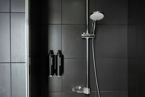 A shower with a handheld showerhead and a wall-mounted faucet, two bottles of shampoo, and a clear plastic shower tray. - The Social Hub Madrid (Madrid)