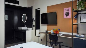 A modern hotel room with a bed, a desk with a laptop, a chair, a TV, a corkboard with a poster, and a plant.