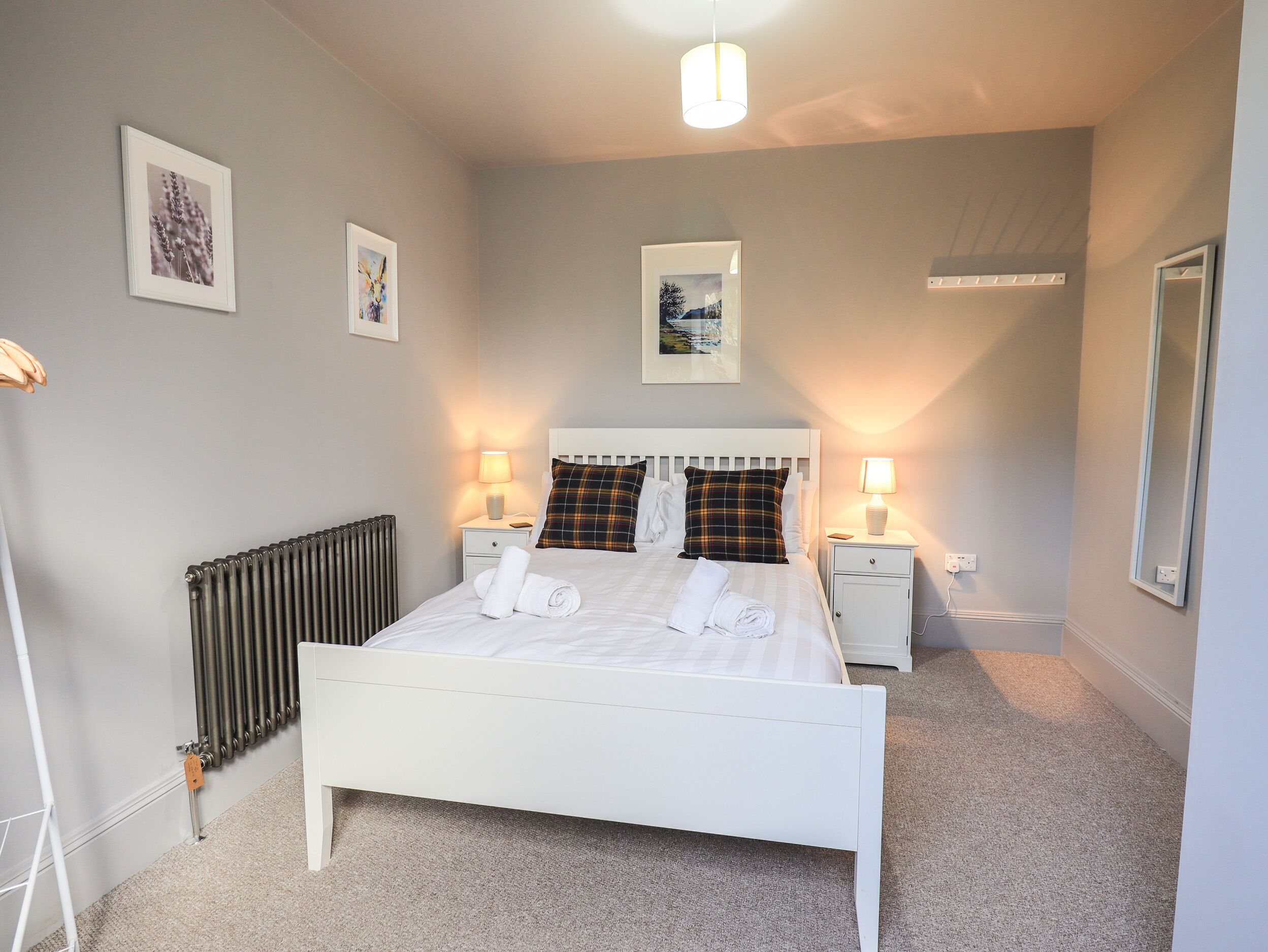 A neatly made bed with white linens and two pillows, a bedside table with a lamp, a radiator, and framed pictures on the wall.