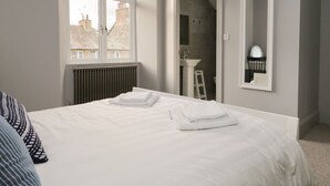 Une chambre avec un grand lit, une fenêtre, un radiateur, une salle de bain visible à travers une porte ouverte et un miroir sur le mur.
