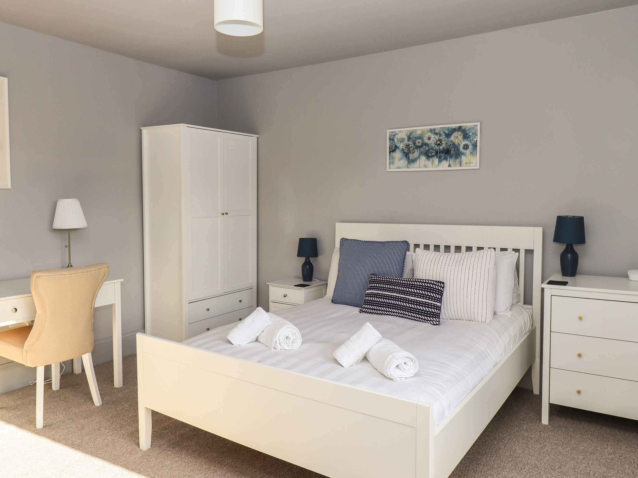 Une chambre à coucher bien rangée, avec un lit, deux tables de chevet, une chaise, une armoire et un tableau accroché au mur.