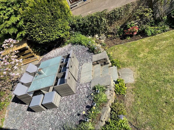 An outdoor seating area with stone tables and chairs, surrounded by a garden with various plants and flowers.
