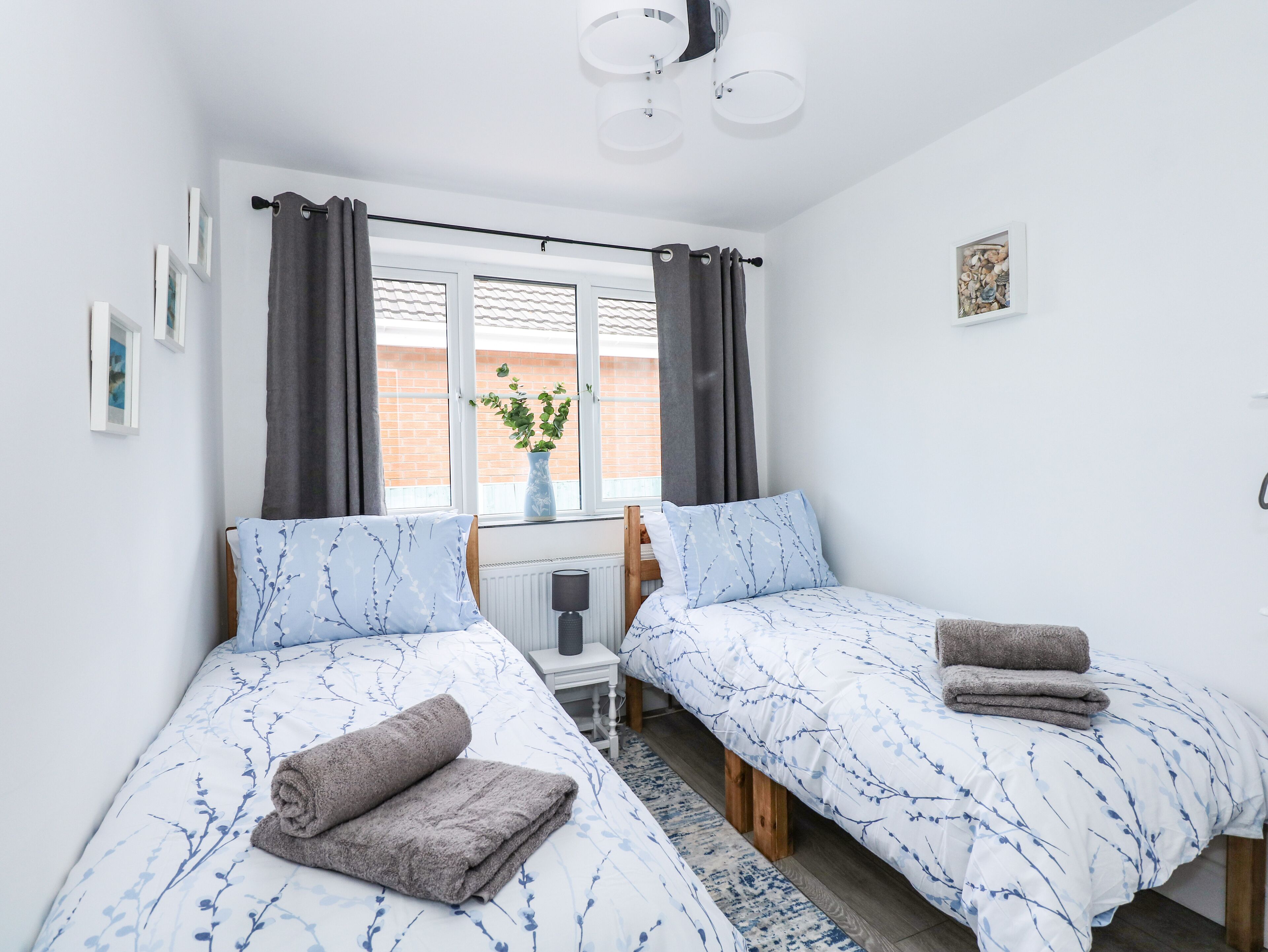 Ein Schlafzimmer mit zwei Betten, einem Fenster mit Vorhängen, einem Heizkörper, einem kleinen Tisch mit Lampe und einer Pflanze auf der Fensterbank.