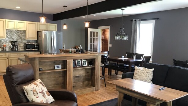A modern kitchen with a central island, wooden dining table, and a black leather sofa.
