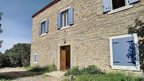 Un edificio de piedra con puerta de madera y contraventanas azules, rodeado de grava y follaje.