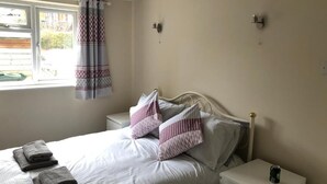Een netjes opgemaakt bed met kussens en opgevouwen handdoeken, een nachtkastje met een blik, en een raam met gordijnen.