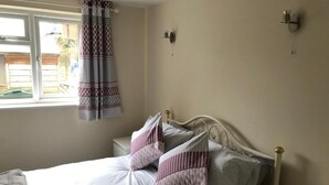 Een net opgemaakt bed met kussens, een nachtkastje met een lamp, en een raam met gordijn.