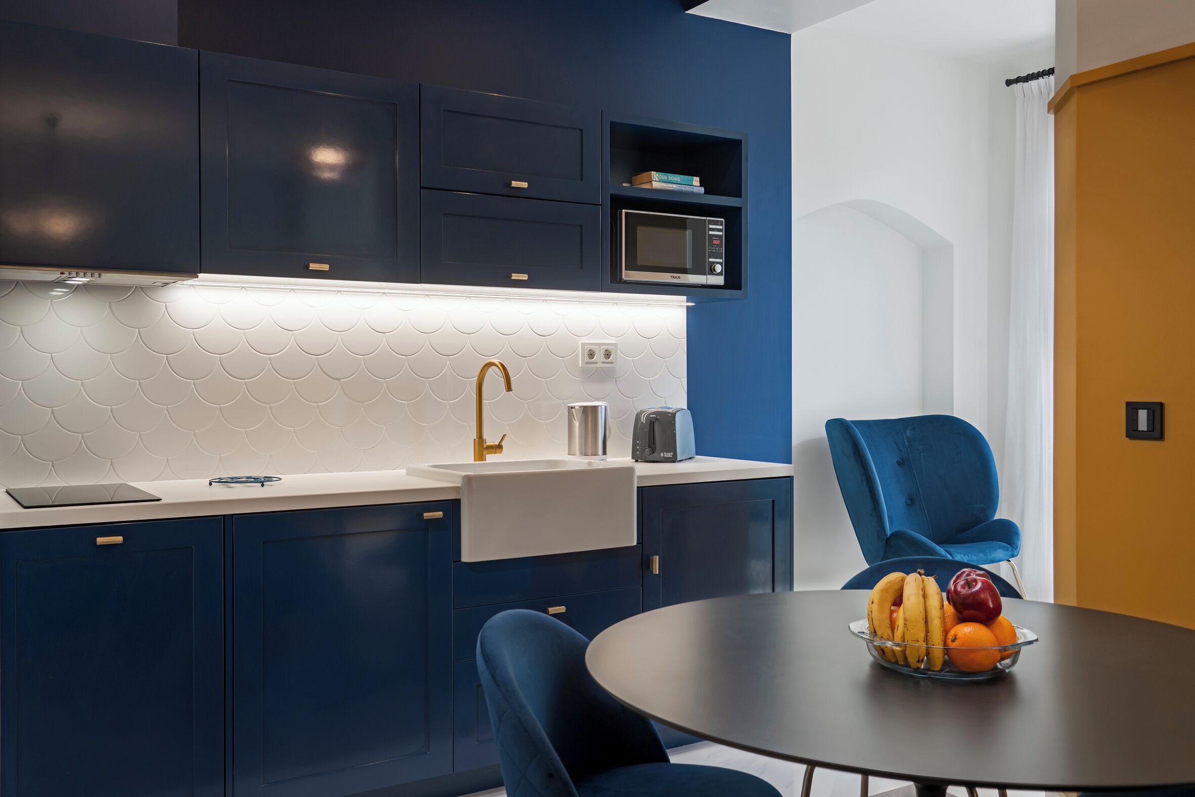 A modern kitchen with dark blue cabinets, a white tiled backsplash, and a microwave. There is a dining table with a bowl of fruit and a blue chair.