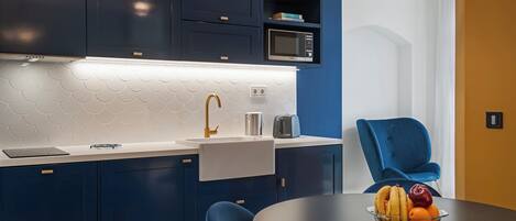 A modern kitchen with dark blue cabinets, a white tiled backsplash, and a microwave. There is a dining table with a bowl of fruit and a blue chair.