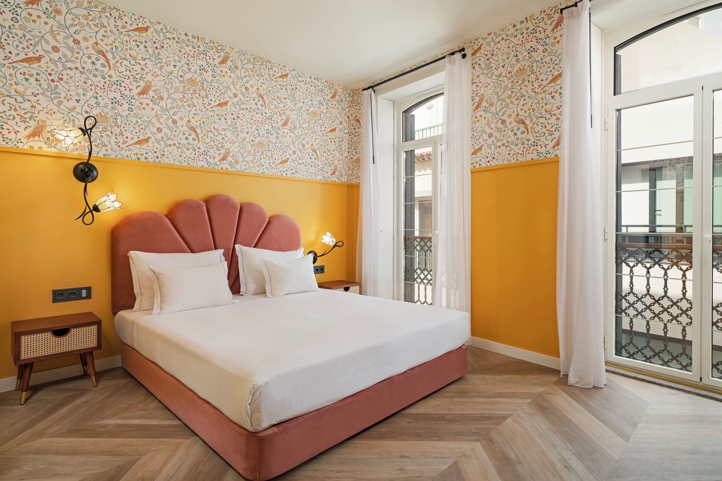 Une chambre dotée d’un grand lit, d’une table de chevet, d’une lampe fixée au mur et d’une fenêtre donnant sur un beau paysage.