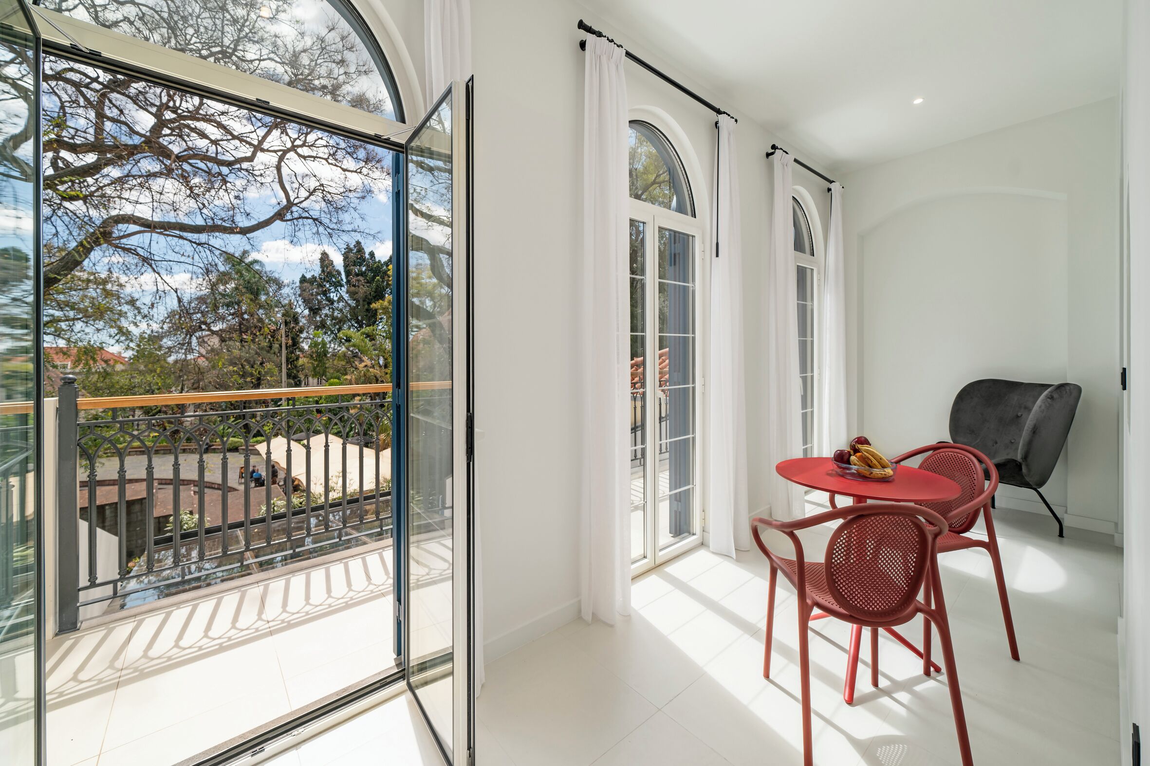 A modern room with a glass door leading to a balcony, a round red table with chairs, and a grey armchair.