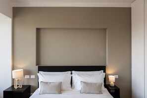 Un lit bien fait, avec du linge de lit blanc et deux oreillers, flanqué de deux tables de chevet sur lesquelles sont posées des lampes, dans une pièce aux murs sobres et dotée d’une grande tête de lit sombre.