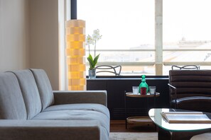 Un salon moderne avec un canapé gris, une table basse en verre et une vue sur la ville grâce à de grandes fenêtres.