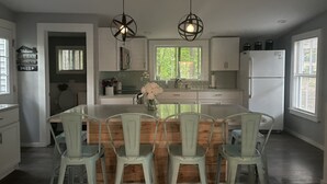A modern kitchen with a central island, bar stools, pendant lighting, and a view of greenery outside. - Cozy 3 bedroom seasonal cottage with very short walk to private community beach (Windham)