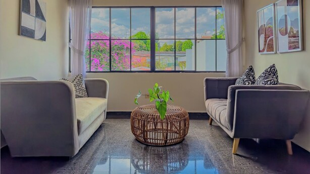 A modern living room with two sofas, a round wicker coffee table, and a ceiling fan. Large windows offer a view of a garden with pink flowers.