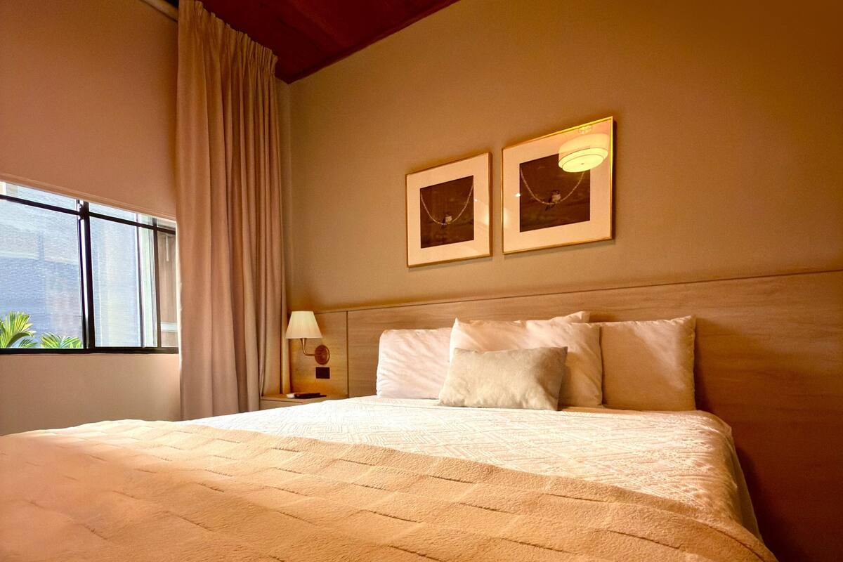 A hotel room with a large bed, a window with curtains, a nightstand with a lamp, and two framed pictures on the wall.