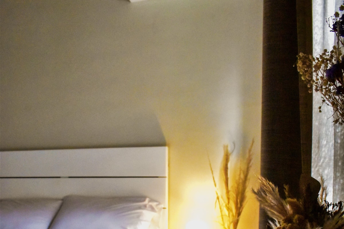 A hotel room with a bed, bedside table, lamp, and a vase with dried flowers.