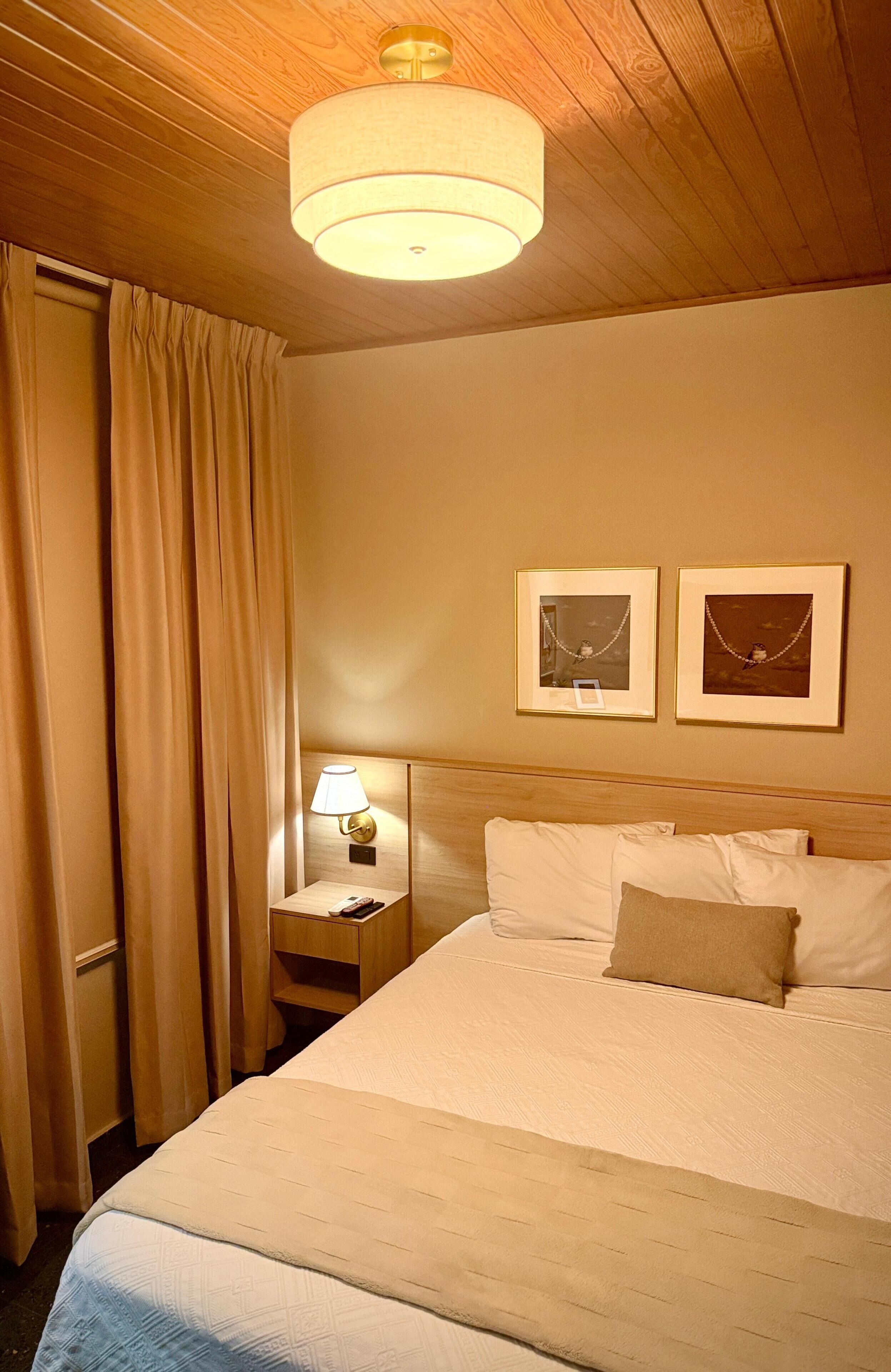 Ein Hotelzimmer mit einem Bett, einem Nachttisch mit Lampe, einem Wandbild und einer Deckenleuchte.