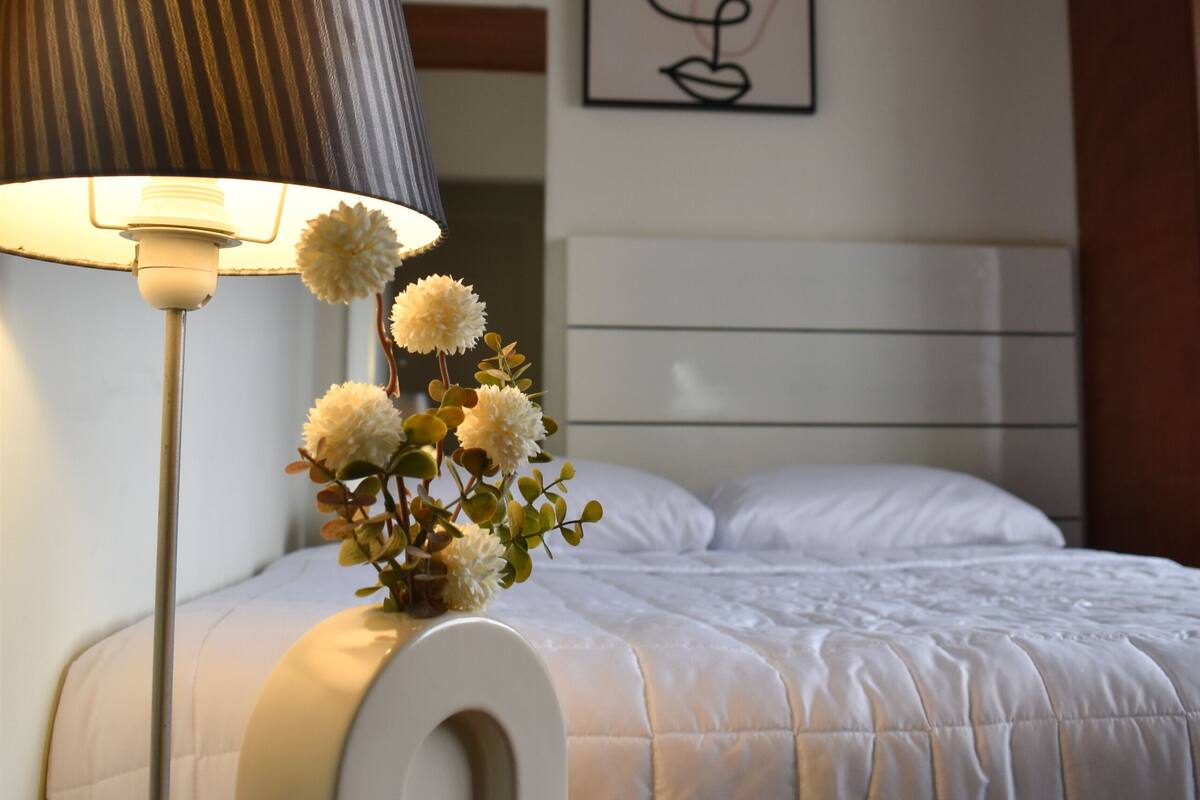 A neatly made bed with a white comforter, a lamp with a striped lampshade, and a small vase with white flowers on a bedside table.