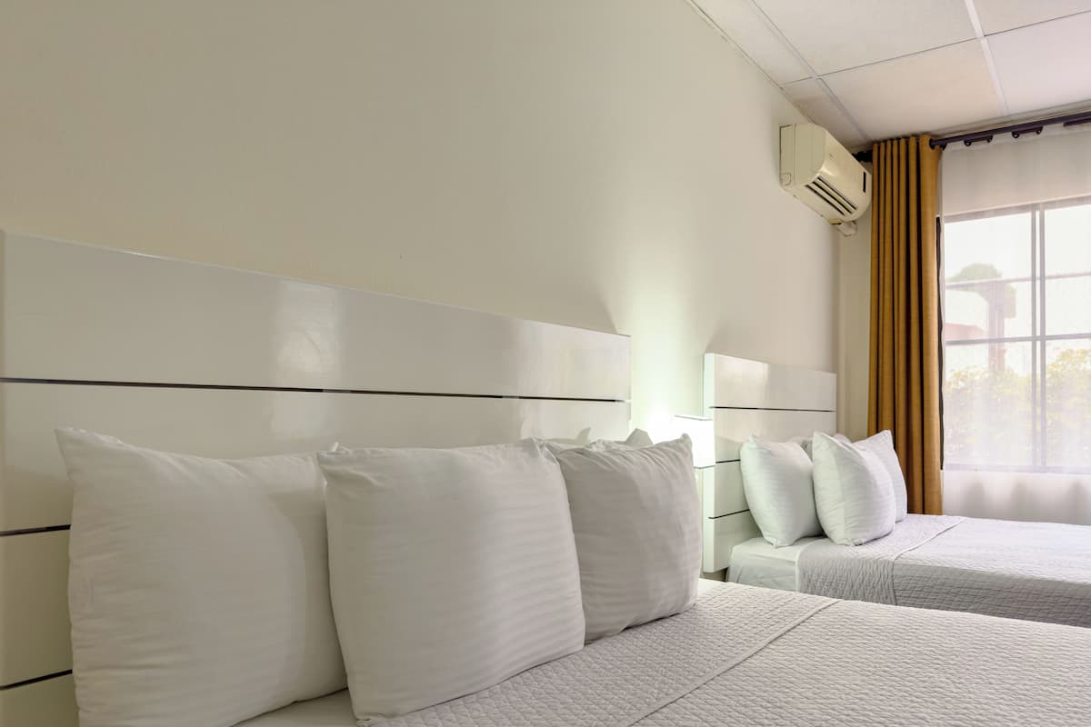 A hotel room with two beds, a headboard, a window with curtains, and an air conditioning unit.