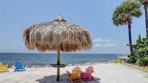 A beach scene with a thatched umbrella, three Adirondack chairs, and a palm tree. - Tiki Themed Condo on Tampa Bay (Tampa)